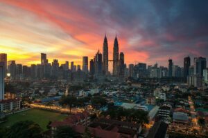 Pemandangan kota Kuala Lumpur ketika senja, dengan langit yang dipenuhi warna - warna merekah seperti merah, oren dan biru. Menara Berkembar Petronas menonjol di antara bangunan - bangunan tinggi. Di bawah, terdapat perkampungan dengan rumah - rumah yang berbeza - beza. Tentang BaoLiba – Mengubah Landskap Pemasaran Influencer di Malaysia: Gambar ini menggambarkan kekayaan dan keanekaragaman Malaysia. BaoLiba boleh membantu mengubah landskap pemasaran influencer di sini, dengan memanfaatkan keindahan kota dan kehidupan tempatan untuk campaigns yang lebih menarik.