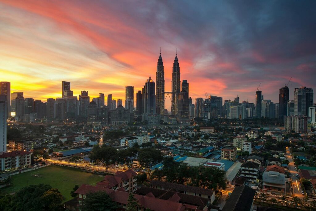 Pemandangan kota Kuala Lumpur ketika senja, dengan langit yang dipenuhi warna - warna merekah seperti merah, oren dan biru. Menara Berkembar Petronas menonjol di antara bangunan - bangunan tinggi. Di bawah, terdapat perkampungan dengan rumah - rumah yang berbeza - beza. Tentang BaoLiba – Mengubah Landskap Pemasaran Influencer di Malaysia: Gambar ini menggambarkan kekayaan dan keanekaragaman Malaysia. BaoLiba boleh membantu mengubah landskap pemasaran influencer di sini, dengan memanfaatkan keindahan kota dan kehidupan tempatan untuk campaigns yang lebih menarik.