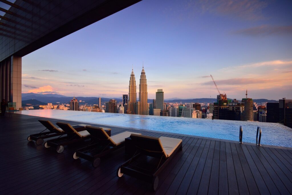 Gambar ini menunjukkan sebuah kolam renang infinity yang terletak di atas bangunan tinggi, dengan pemandangan menakjubkan bandaraya. Di latar belakang, terdapat menara - menara megah termasuk Menara Petronas yang terkenal. Di tepi kolam, terdapat beberapa kerusi berkemas untuk berehat.
